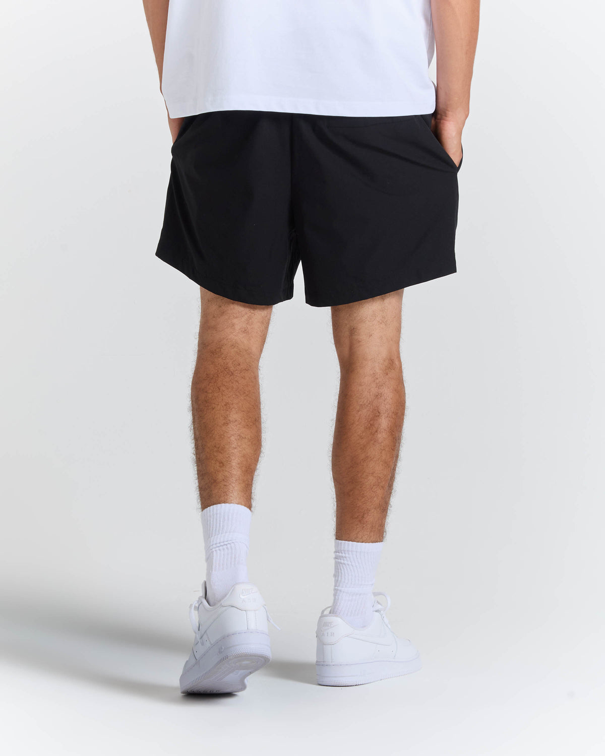 A person shown from the back wearing black athletic shorts, a white t-shirt, white socks, and white sneakers against a light gray studio background.