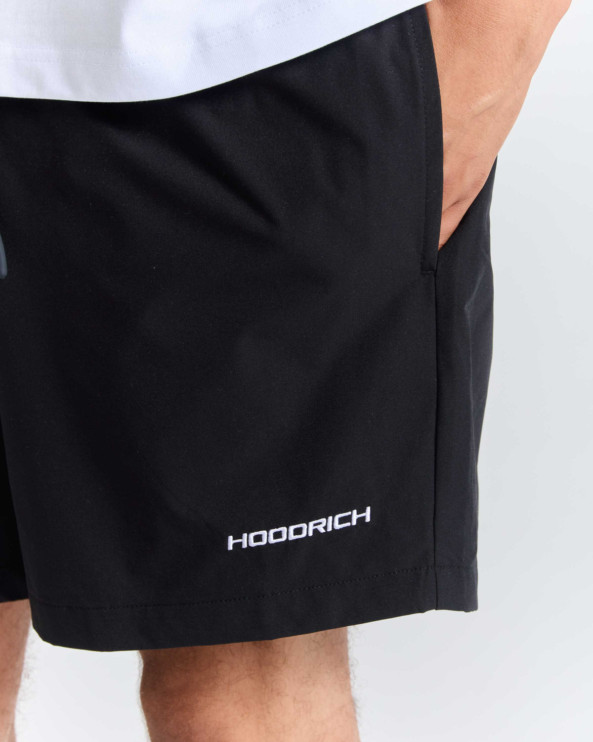 Close-up of a person wearing black athletic shorts with a white HOODRICH logo near the hem, against a light gray background.