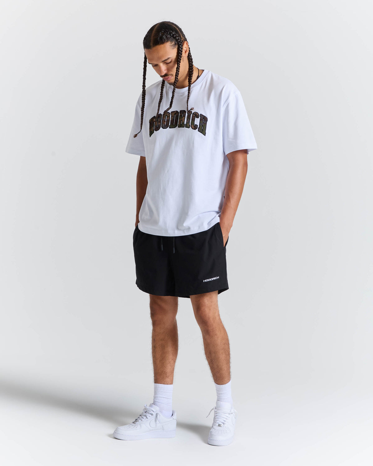 A man modeling a white t-shirt with black 'HOODRICH' text and black shorts, paired with white socks and white sneakers, against a light gray studio background.