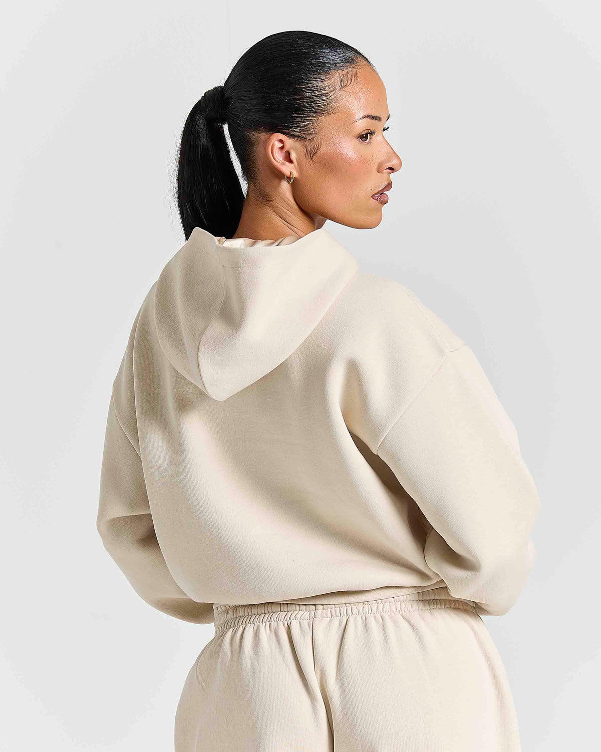 A woman with her hair in a ponytail is shown from the back wearing a cream-colored oversized hoodie and matching sweatpants against a light gray studio background.