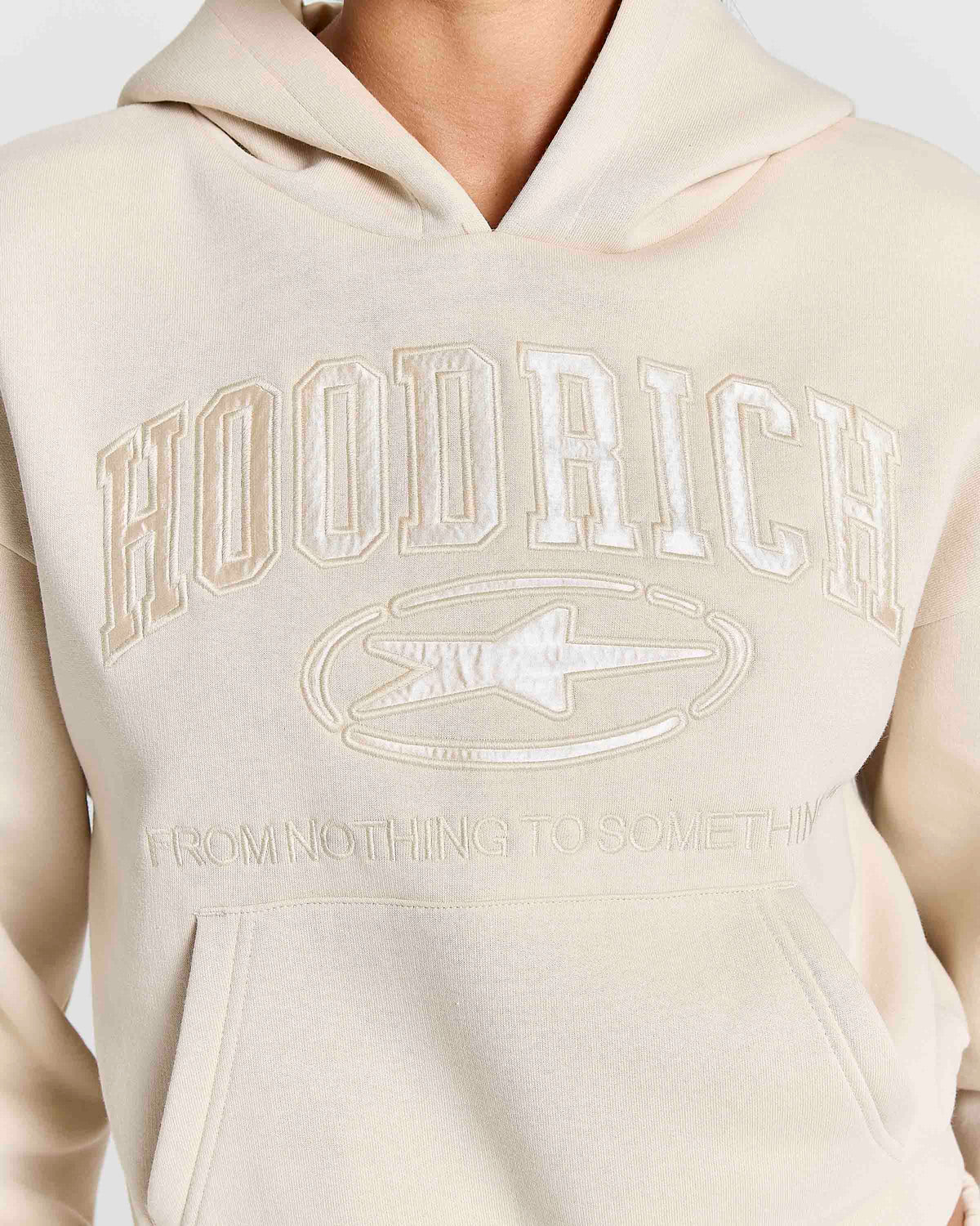 A close-up of a person wearing a cream-colored hoodie with tonal embroidered lettering that reads 'HOODRICH' and a star logo on the chest.