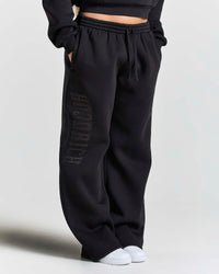 Black wide-leg sweatpants with a tonal vertical 'HOODRICH' logo on the left leg, shown on a model wearing white sneakers against a light gray background.