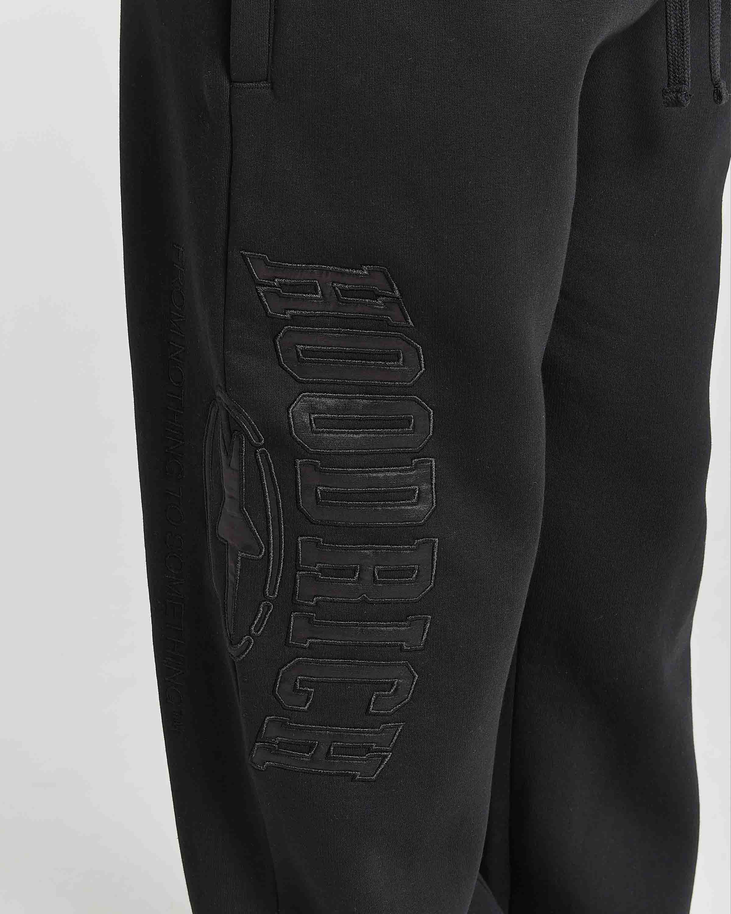 Close-up of black sweatpants featuring a large tonal black-on-black 'HOODRICH' logo and circular emblem on the left leg, shown against a light gray background.