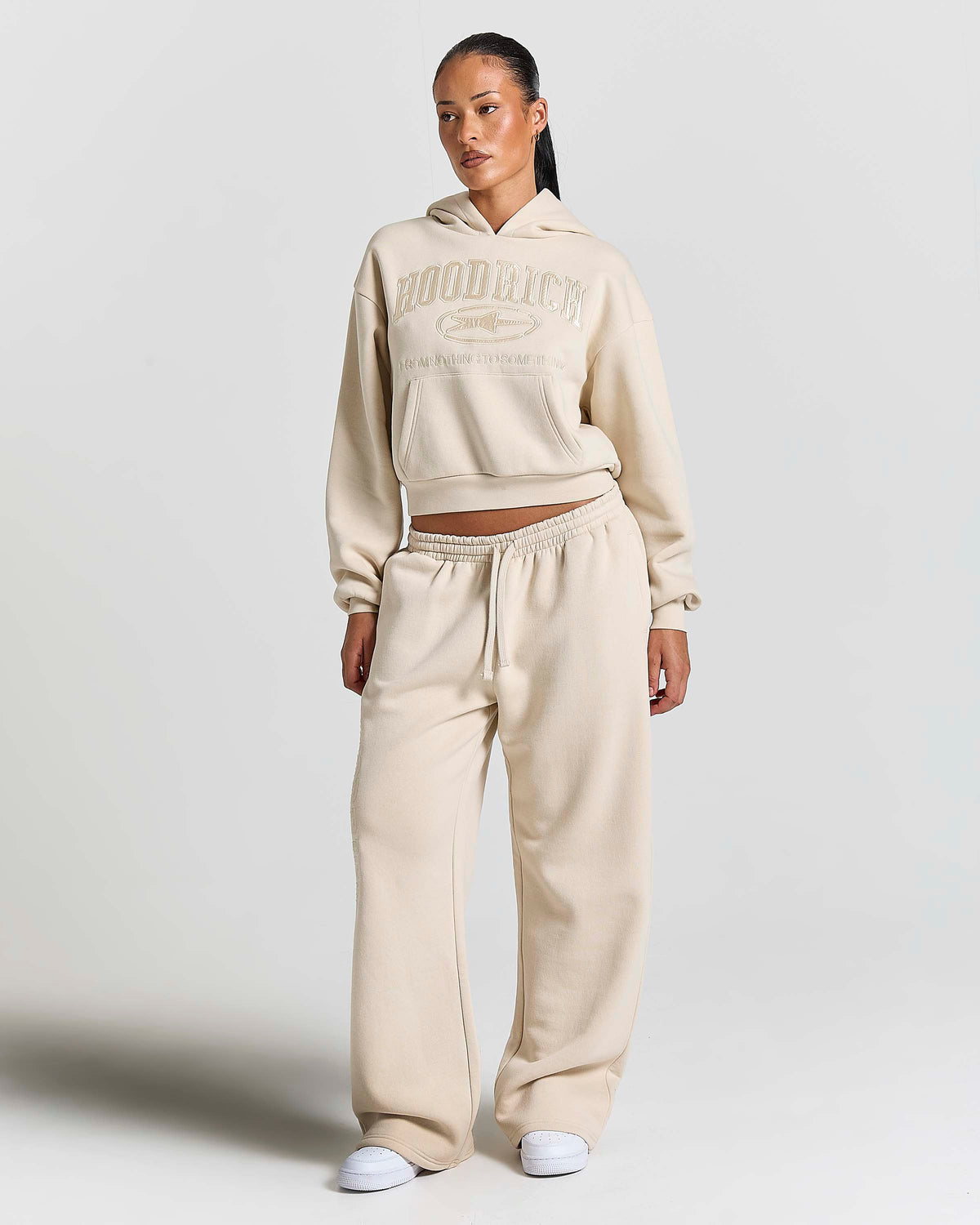 A model wearing a matching beige cropped hoodie and wide-leg sweatpants set with white sneakers, standing against a light gray studio background.
