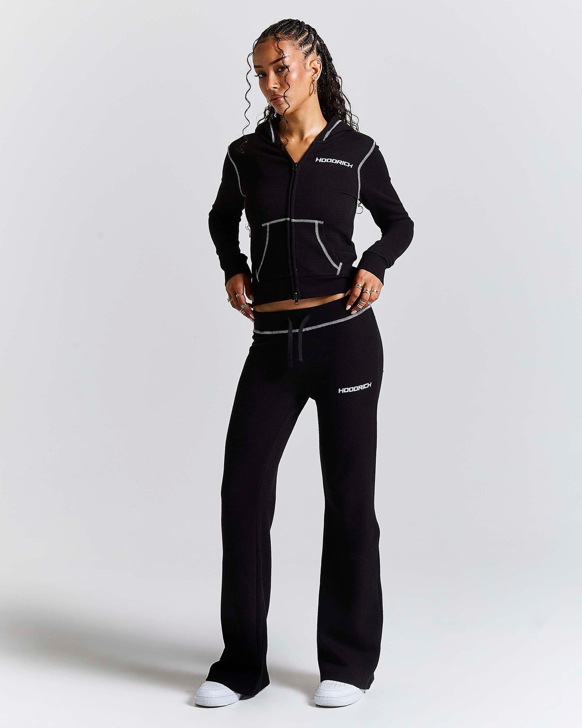 A woman modeling a black athletic tracksuit with white contrast stitching and white logo text on the chest and thigh, photographed against a light gray studio background.