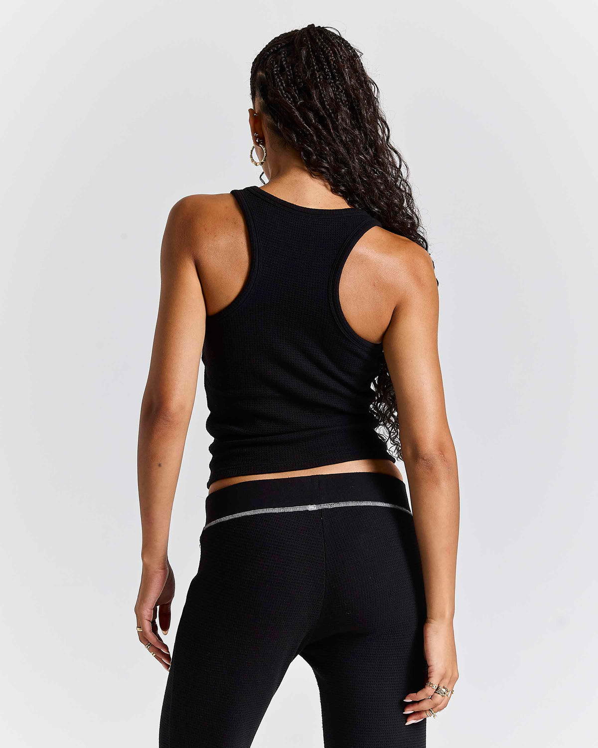 Rear view of a woman wearing a black ribbed athletic tank top and black leggings with a thin silver stripe detail on the waistband, set against a light gray studio background.