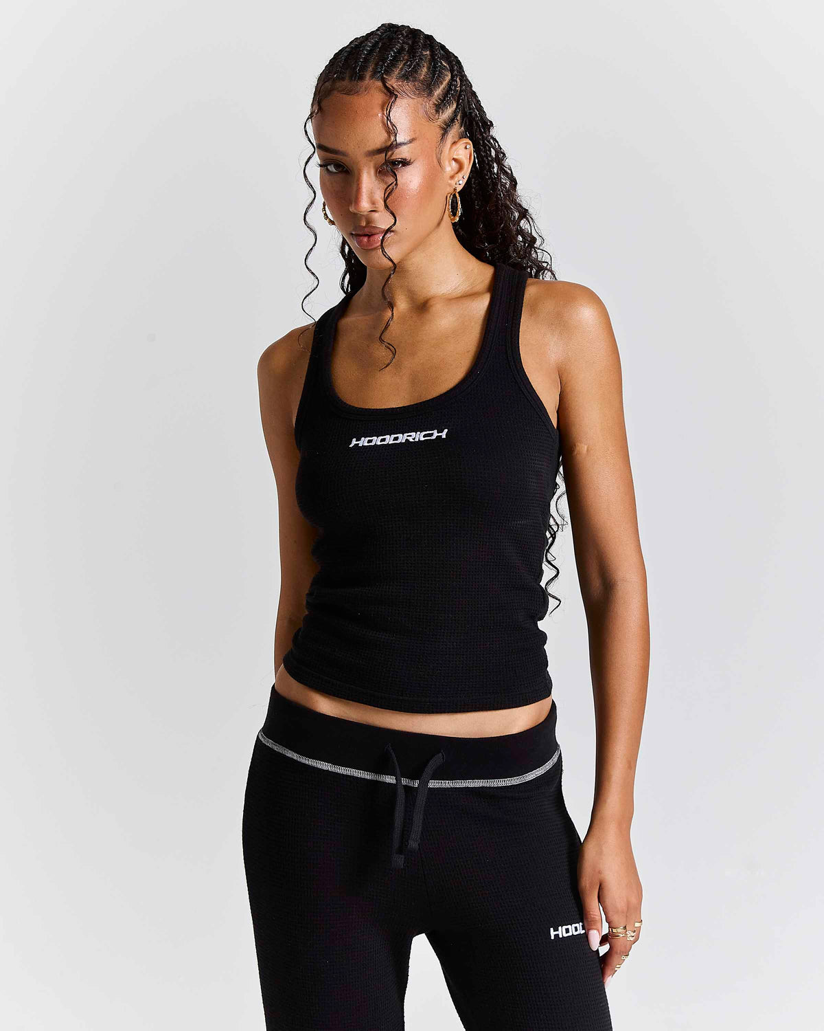 A woman modeling a black ribbed athletic tank top and matching black joggers with white accents, featuring the brand name 'HOODRICH' on the chest and upper thigh.
