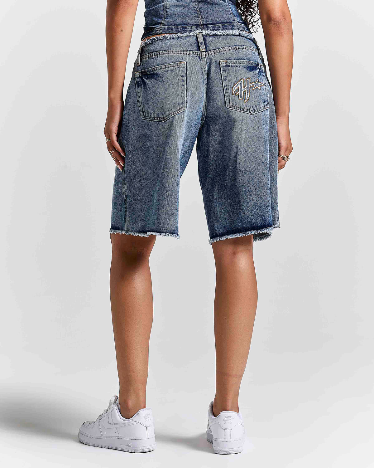 A person shown from the back wearing medium-wash blue denim shorts with a raw frayed hem, paired with white sneakers against a light gray studio background.