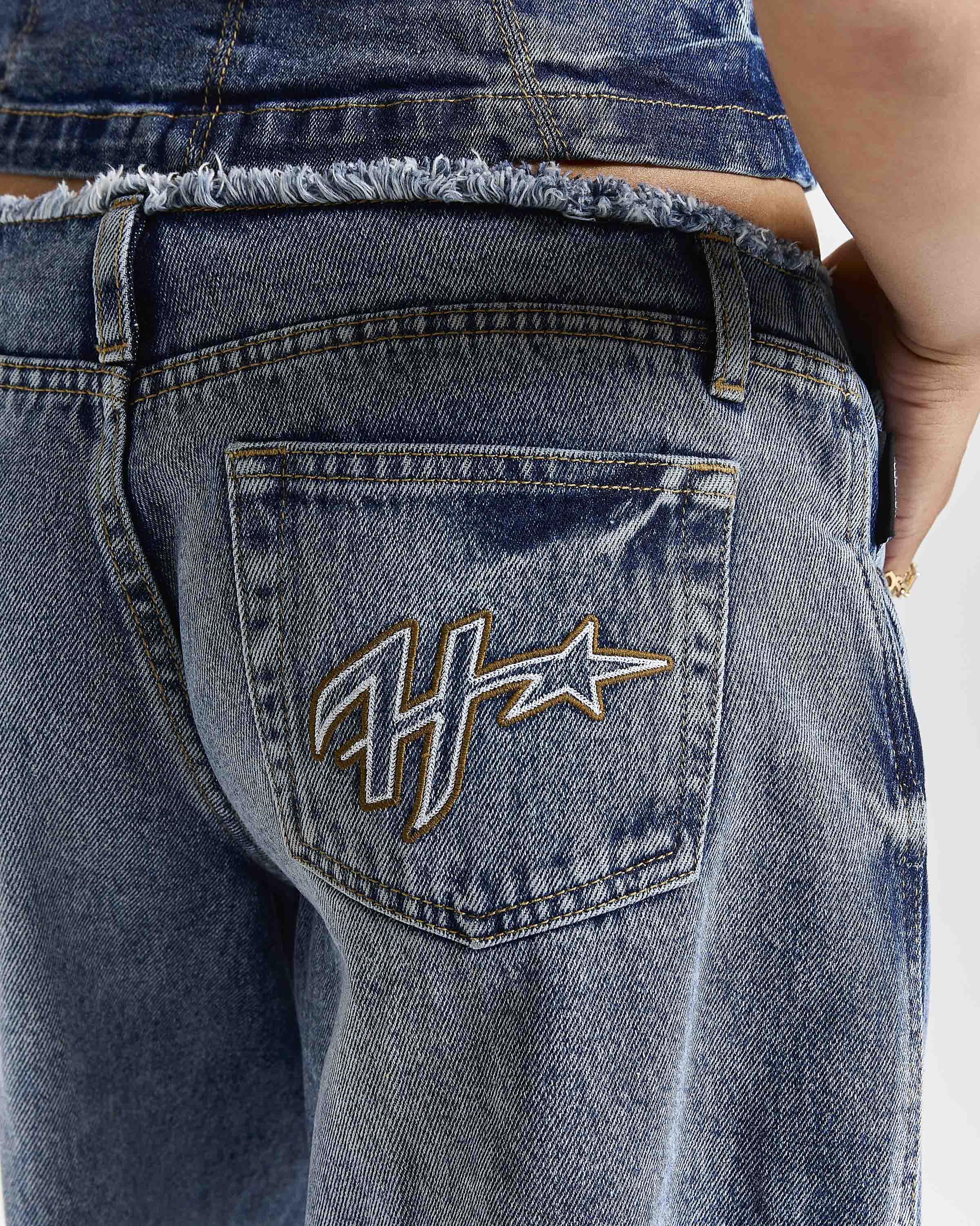 Close-up back view of a person wearing blue denim jeans with a frayed waistband and a large embroidered monogram logo on the back pocket.