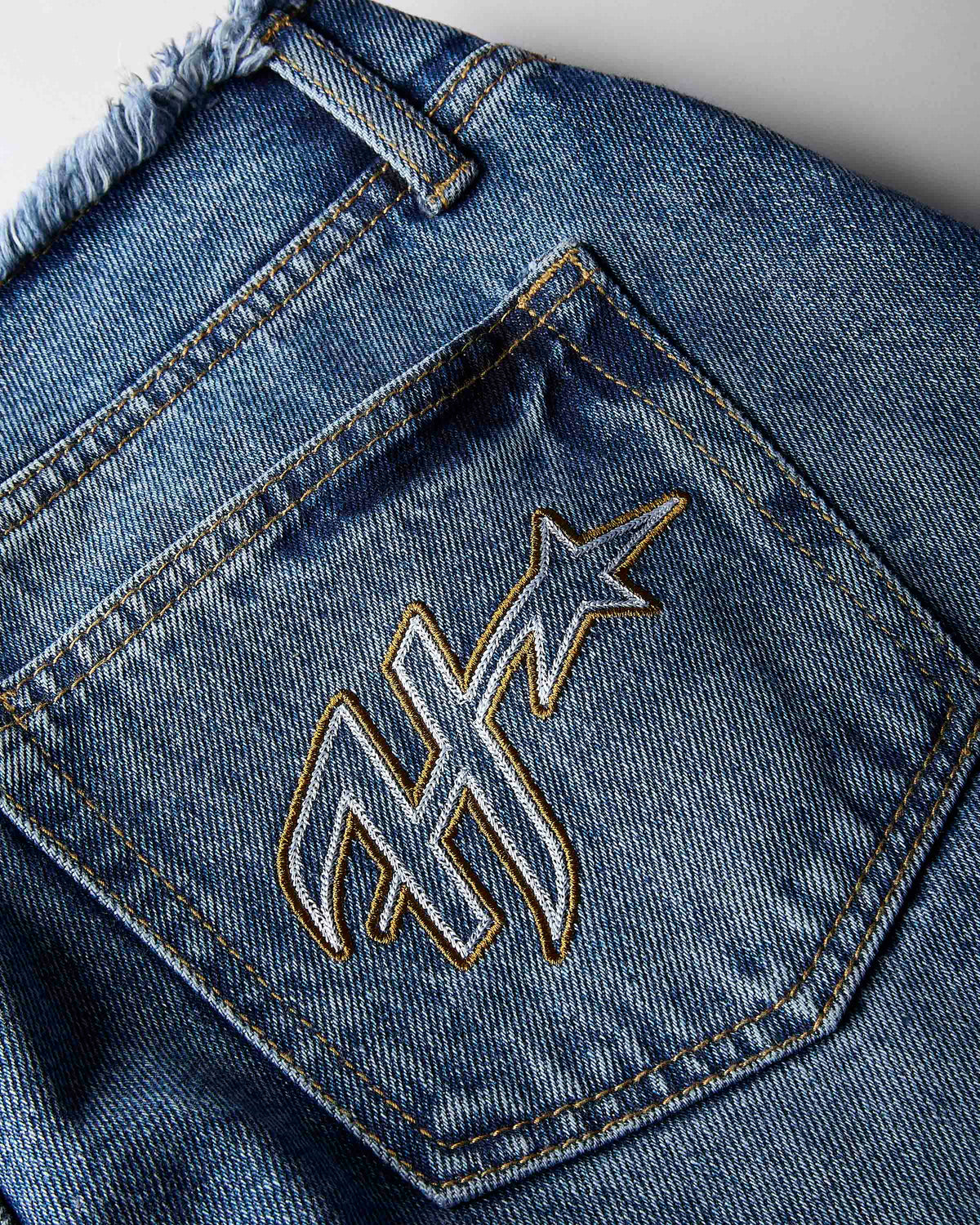 Close-up of the back pocket of blue denim jeans with a large embroidered monogram logo outlined in gold thread.