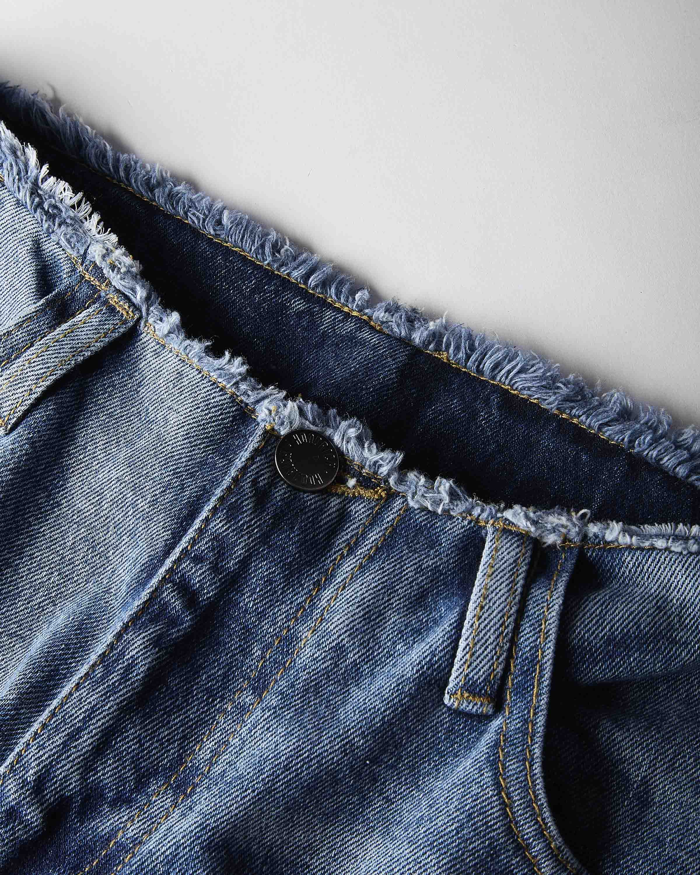 Close-up of a pair of blue denim jeans showing the raw, frayed waistband, a single button closure, and belt loops against a light gray background.