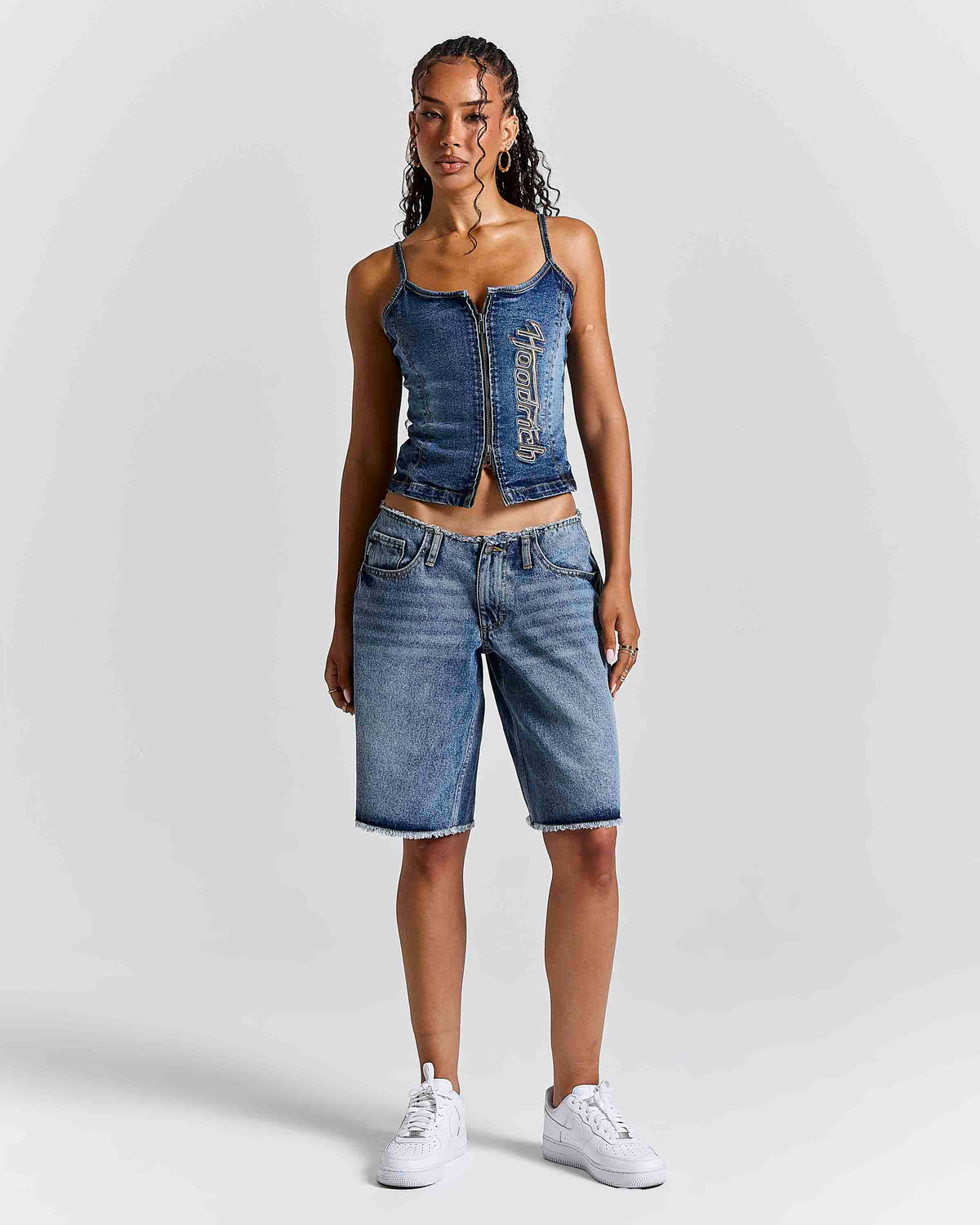 A model stands against a light gray studio background wearing a blue denim crop top with a front zipper and vertical text detail, matching blue denim shorts, and white sneakers.