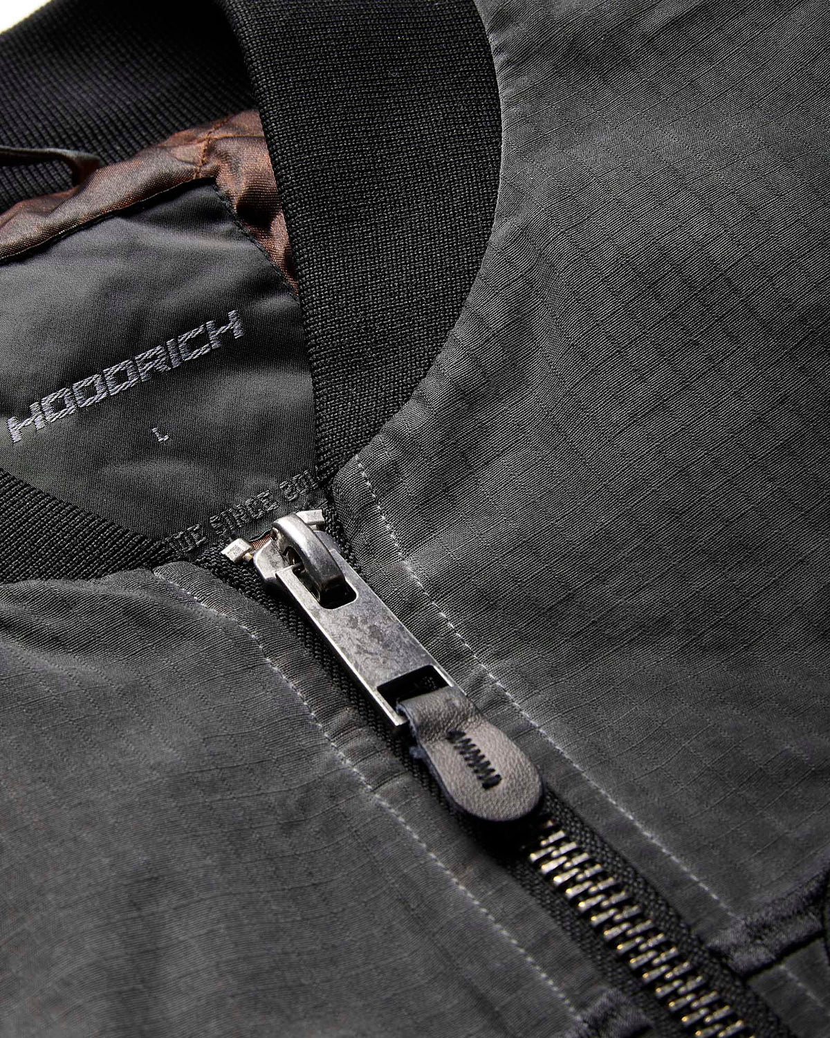 Close-up of a dark gray zippered jacket with a black ribbed collar and a silver metal zipper pull. The brand name 'HOODRICH' is printed on the inner neck lining.