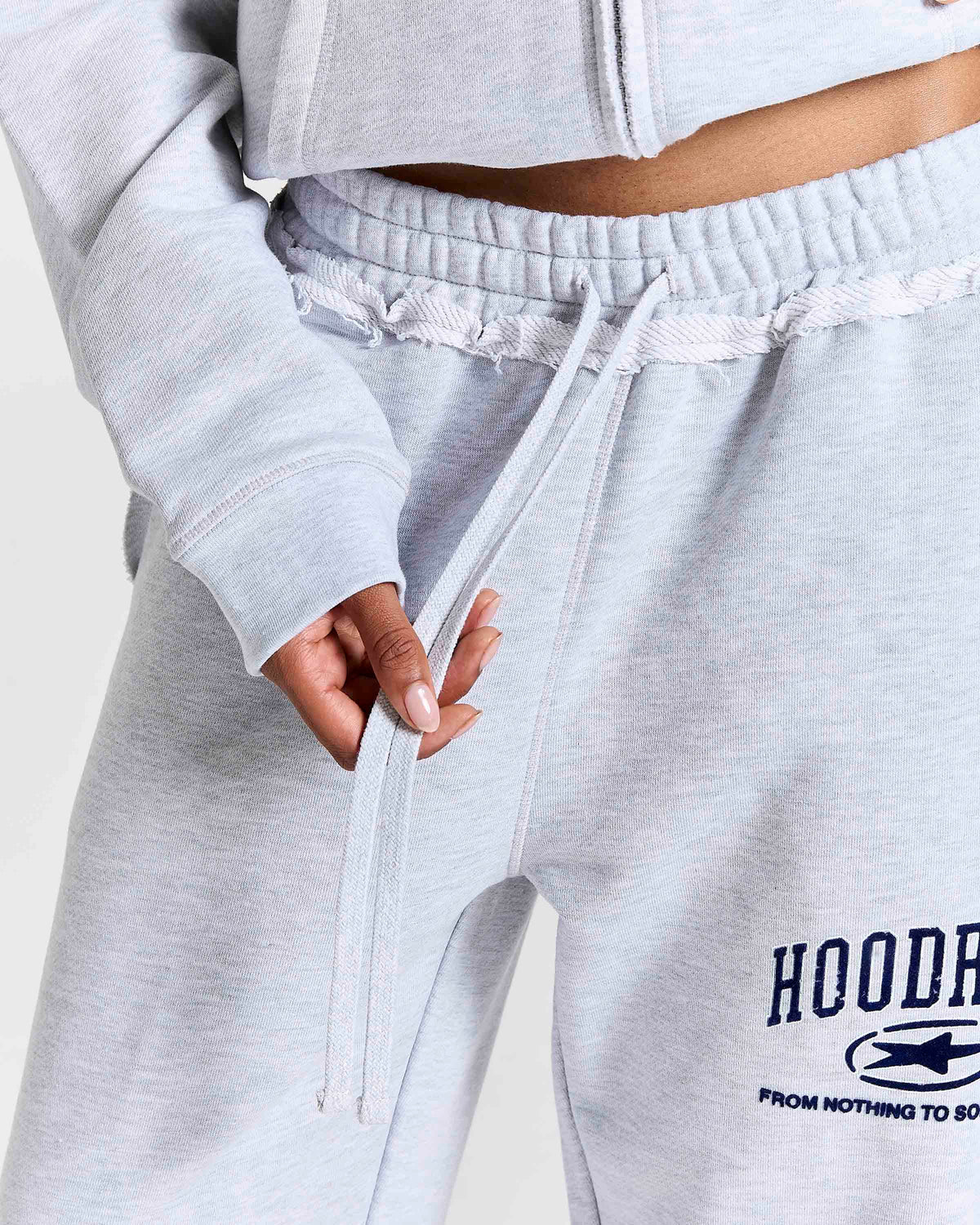 Close-up of a person wearing light heather gray sweatpants with a drawstring waist and a Navy graphic print on the left leg that reads 'HOODRICH' with a small logo and 'FROM NOTHING TO SOMETHING'.