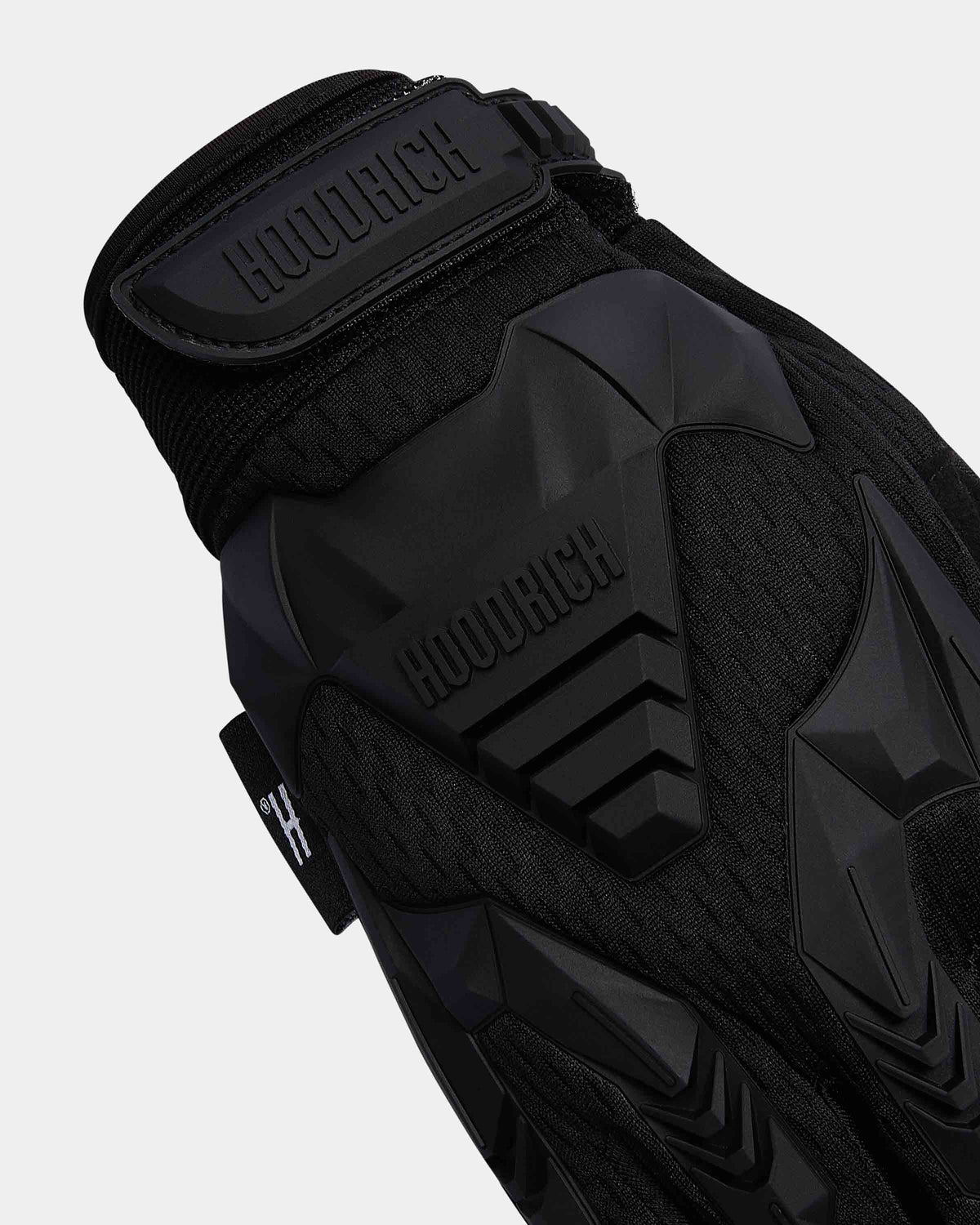 Close-up of a black HOODRICH protective glove showing the wrist strap, textured knit fabric, and molded black protective plates on a light gray background.