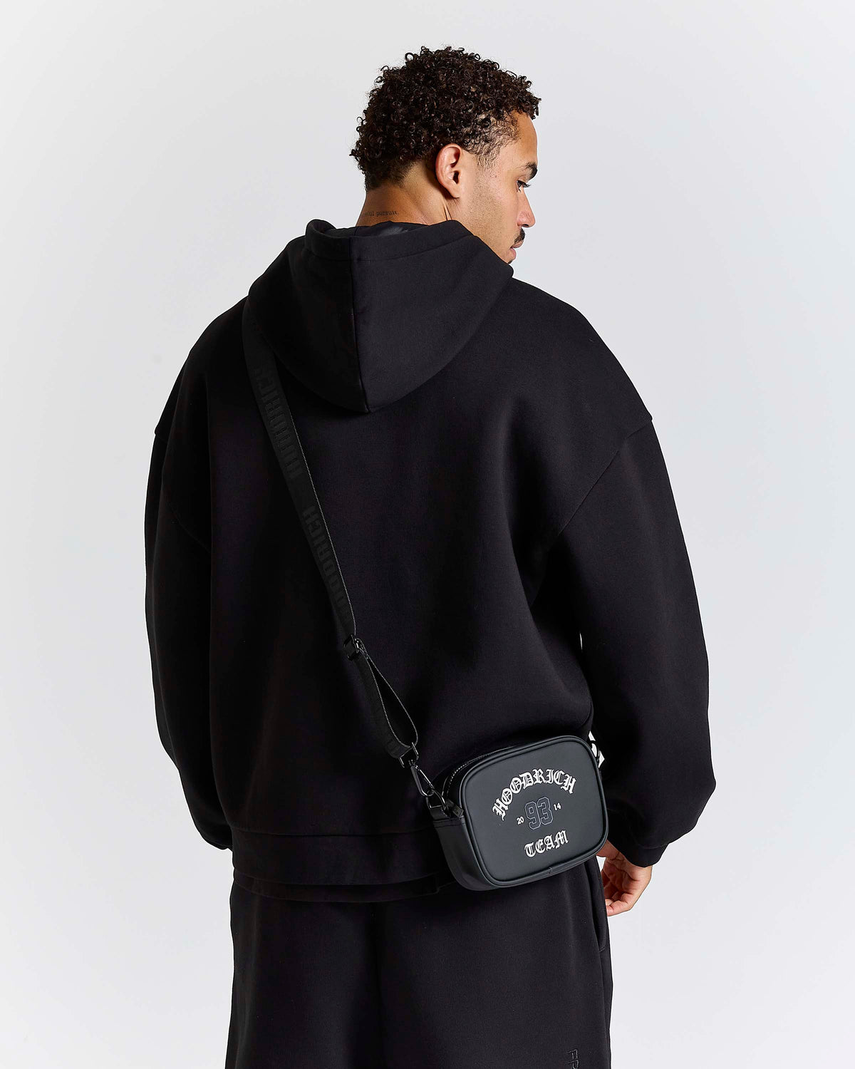 A man wearing a black hoodie and black pants, shown from the back, wearing a black crossbody bag with white text that reads 'HOODRICH' and 'TEAM'.