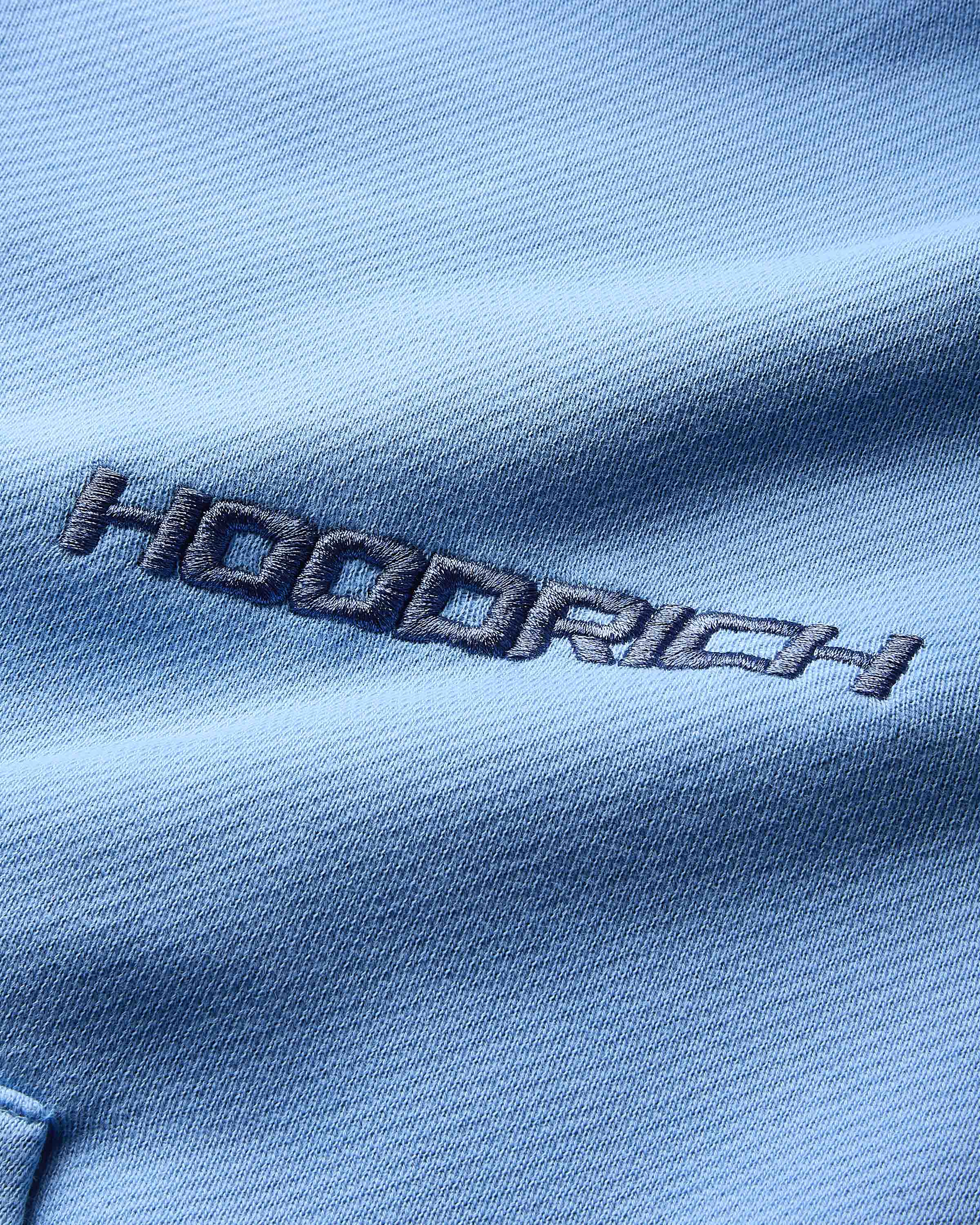 Close-up of the dark blue Hoodrich logo embroidered on the blue fabric of the hoodie.