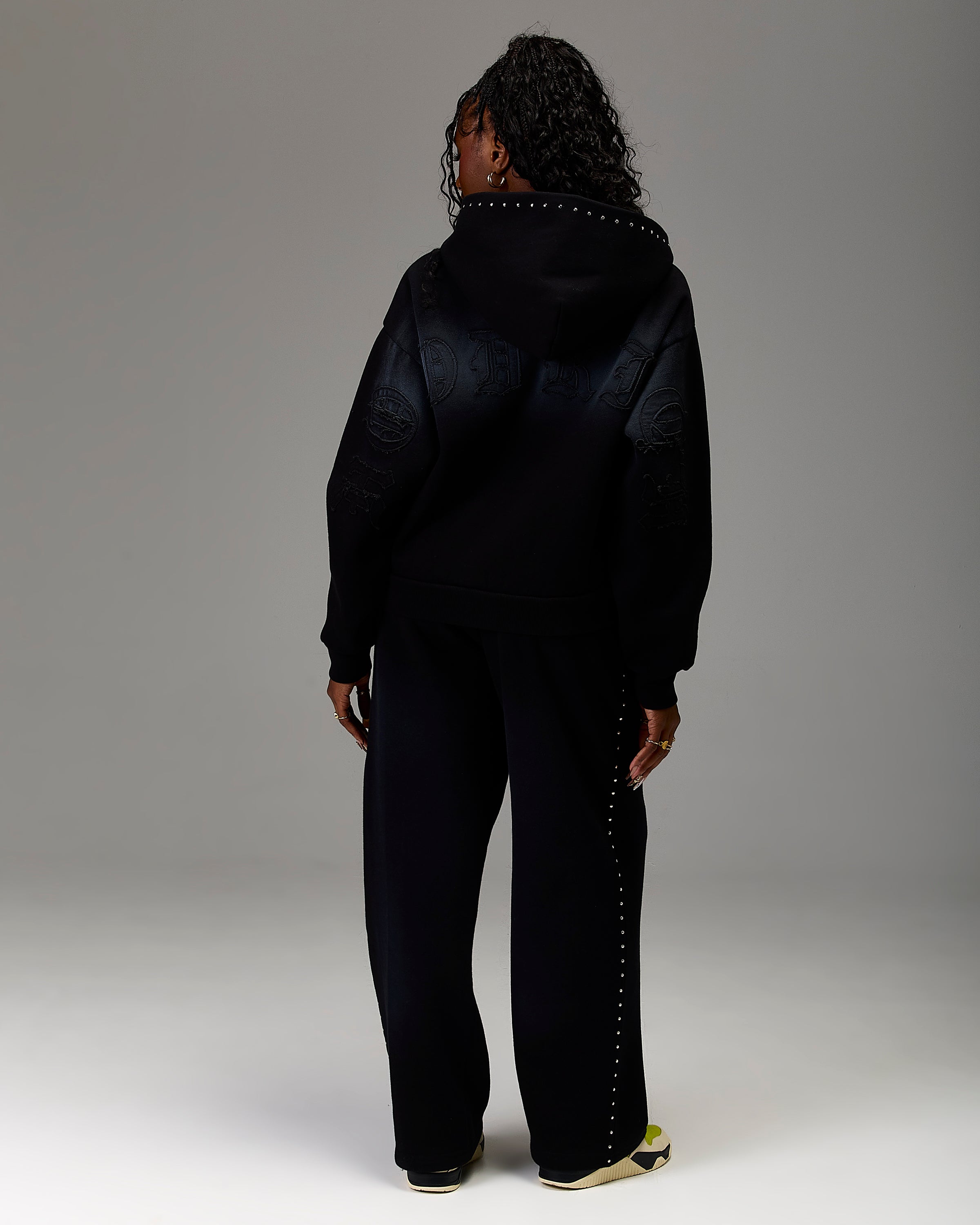 A model viewed from the back wearing a black hooded jacket with tonal embroidered details and matching black wide-leg pants with silver stud accents down the side, set against a neutral gray studio background.