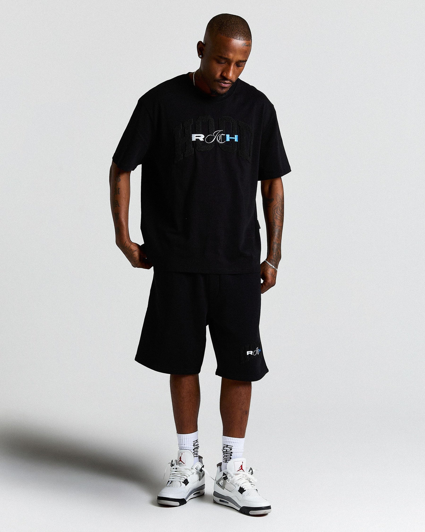 Man wearing a black t-shirt and shorts with a logo, standing against a plain background