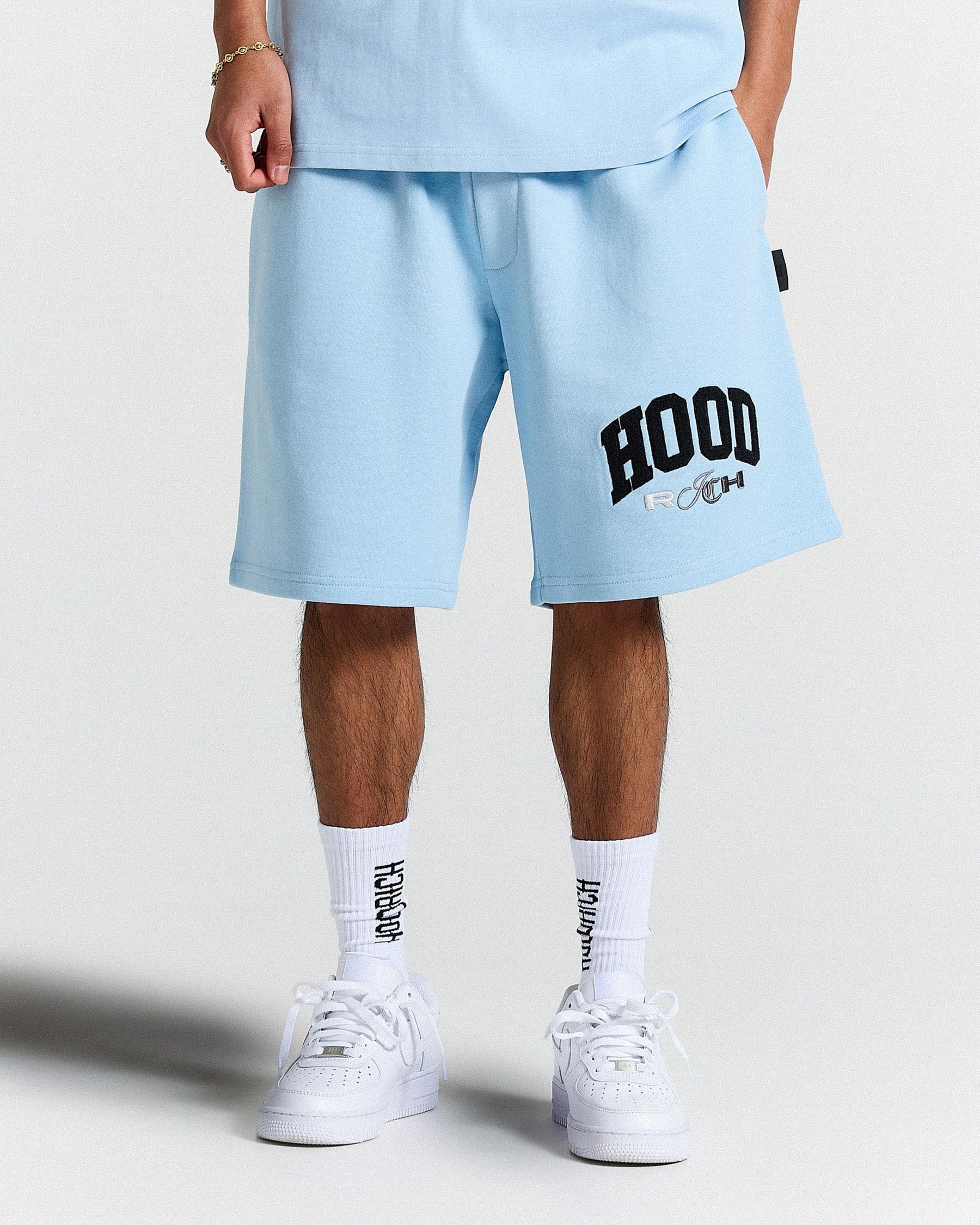 Person wearing light blue shorts with 'HOODRICH' print, white socks, and white sneakers on a light grey background