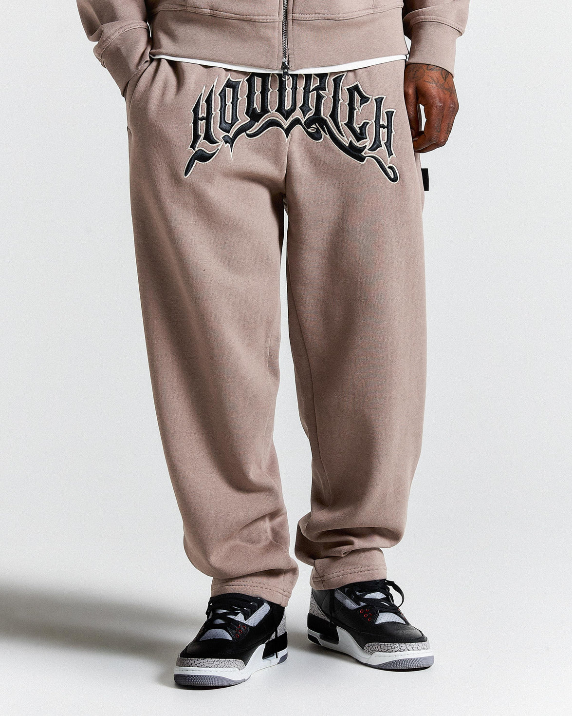 Brown sweatpants with a brand logo on the back, worn by a person, on a white background.