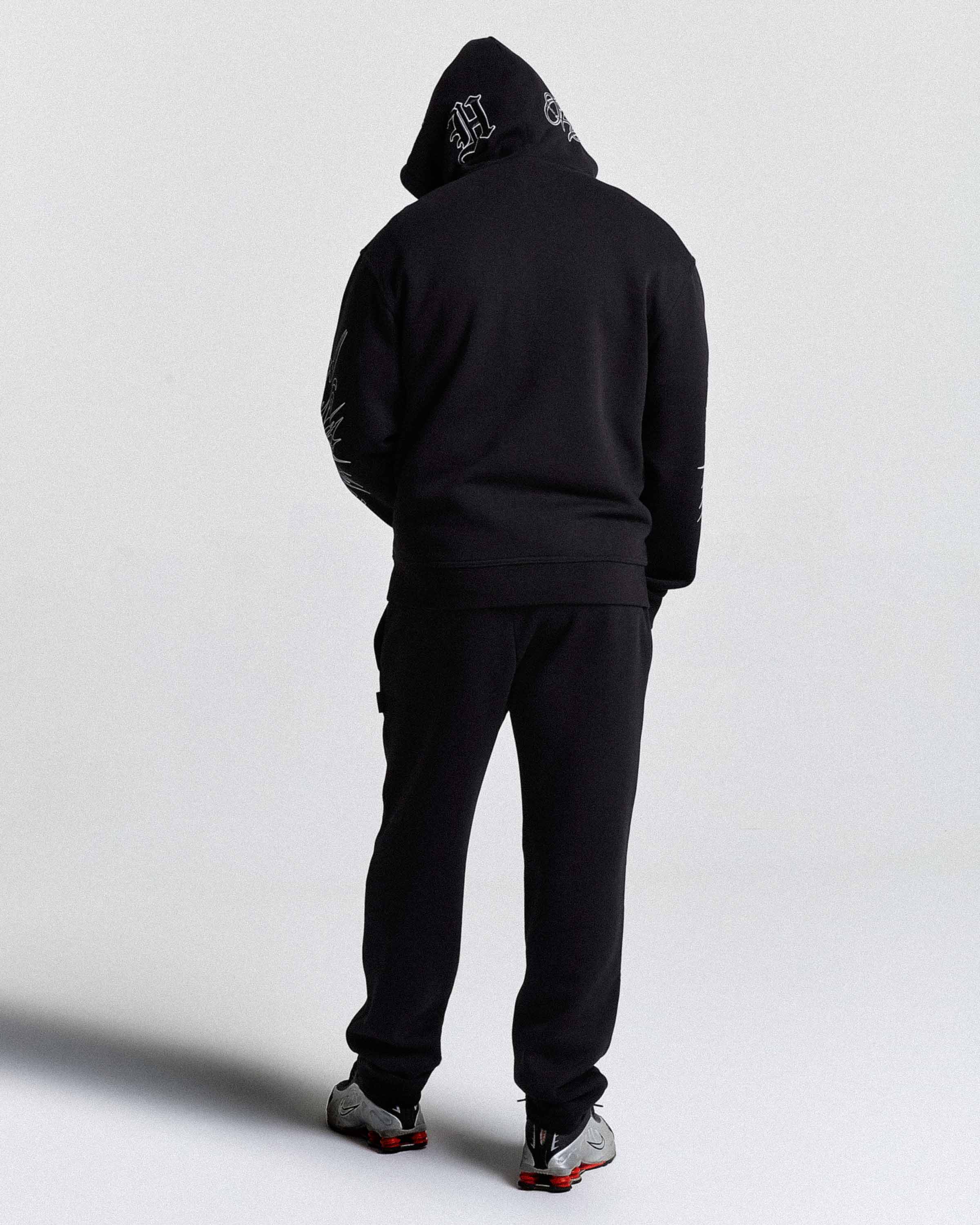 Person wearing a black hoodie and pants with a logo on the hood against a white background