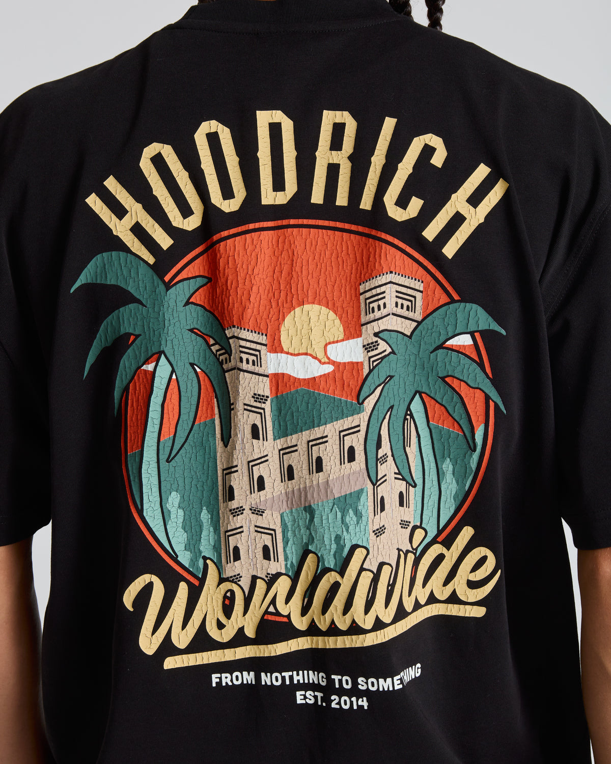 Close-up of the back of a black t-shirt featuring a large graphic print with the words 'HOODRICH Worldwide' in beige, a red sunset, green palm trees, and a beige building.