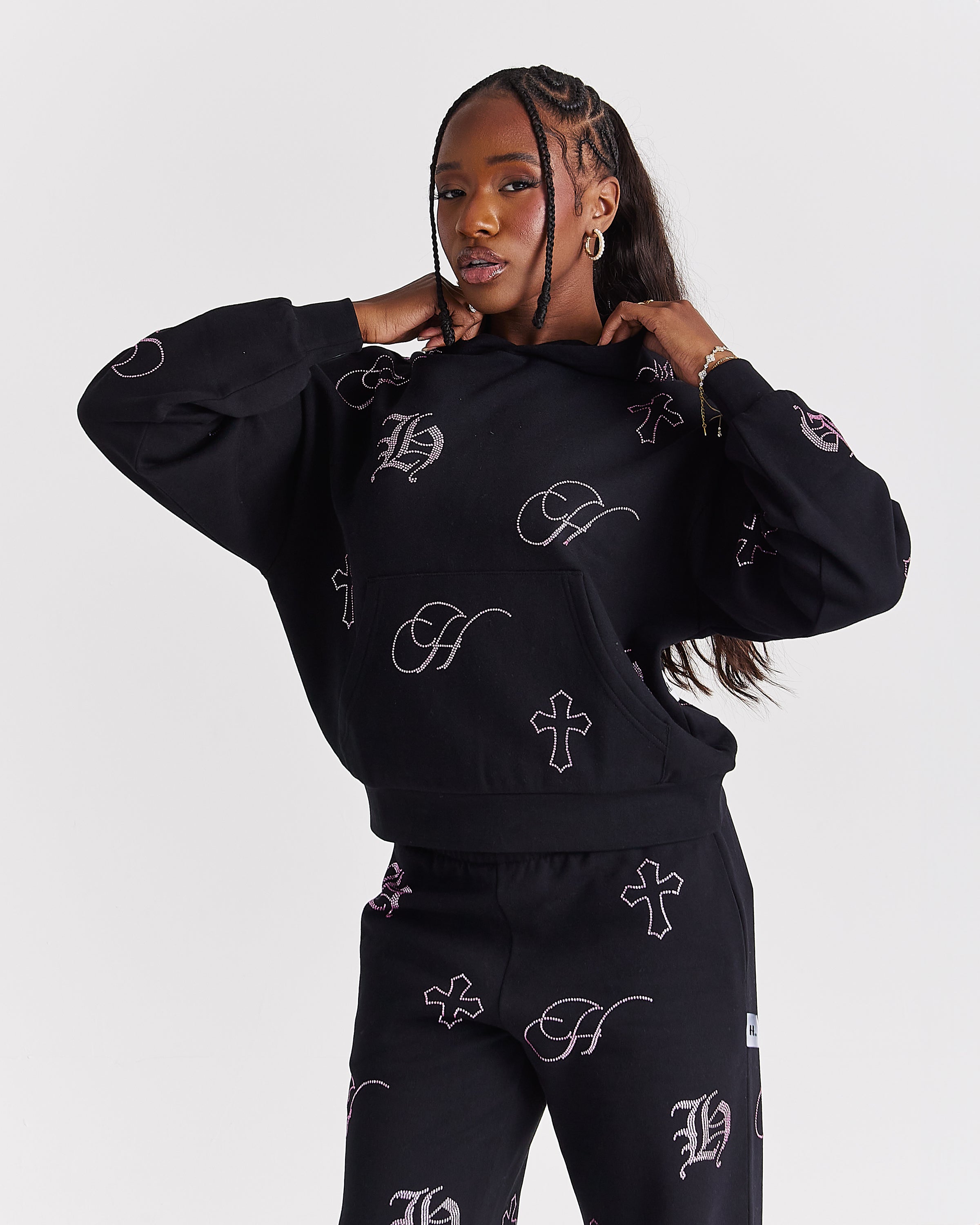 A model wearing a black matching sweatshirt and sweatpants set with an allover white Gothic letter 'H' and cross pattern, posed against a light gray studio background.