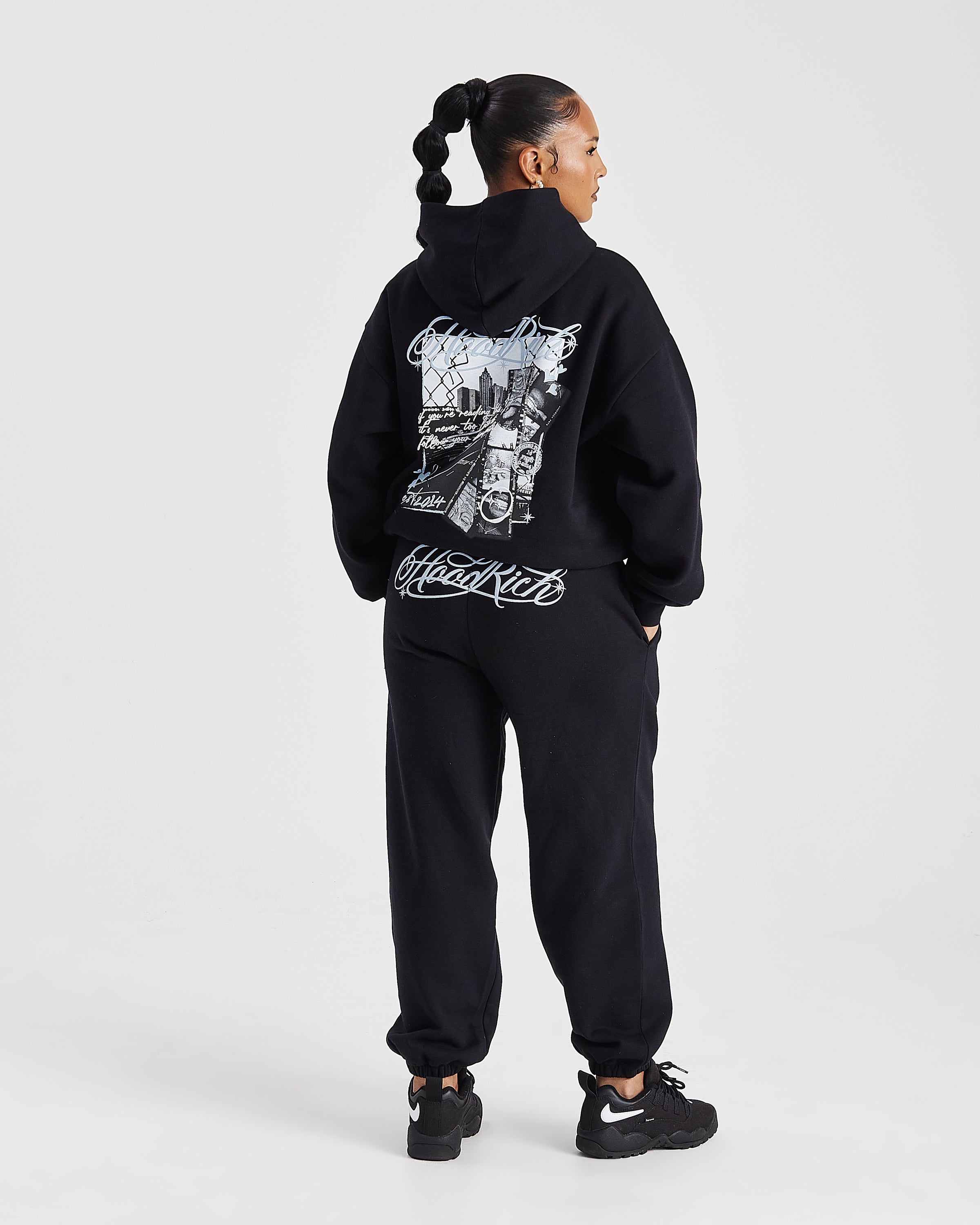 A model shown from the back wearing a black hoodie and sweatpants set with white graphic prints, including a motorcycle illustration on the back of the hoodie and 'Hoodrich' script on the back of the pants, against a light gray studio background.