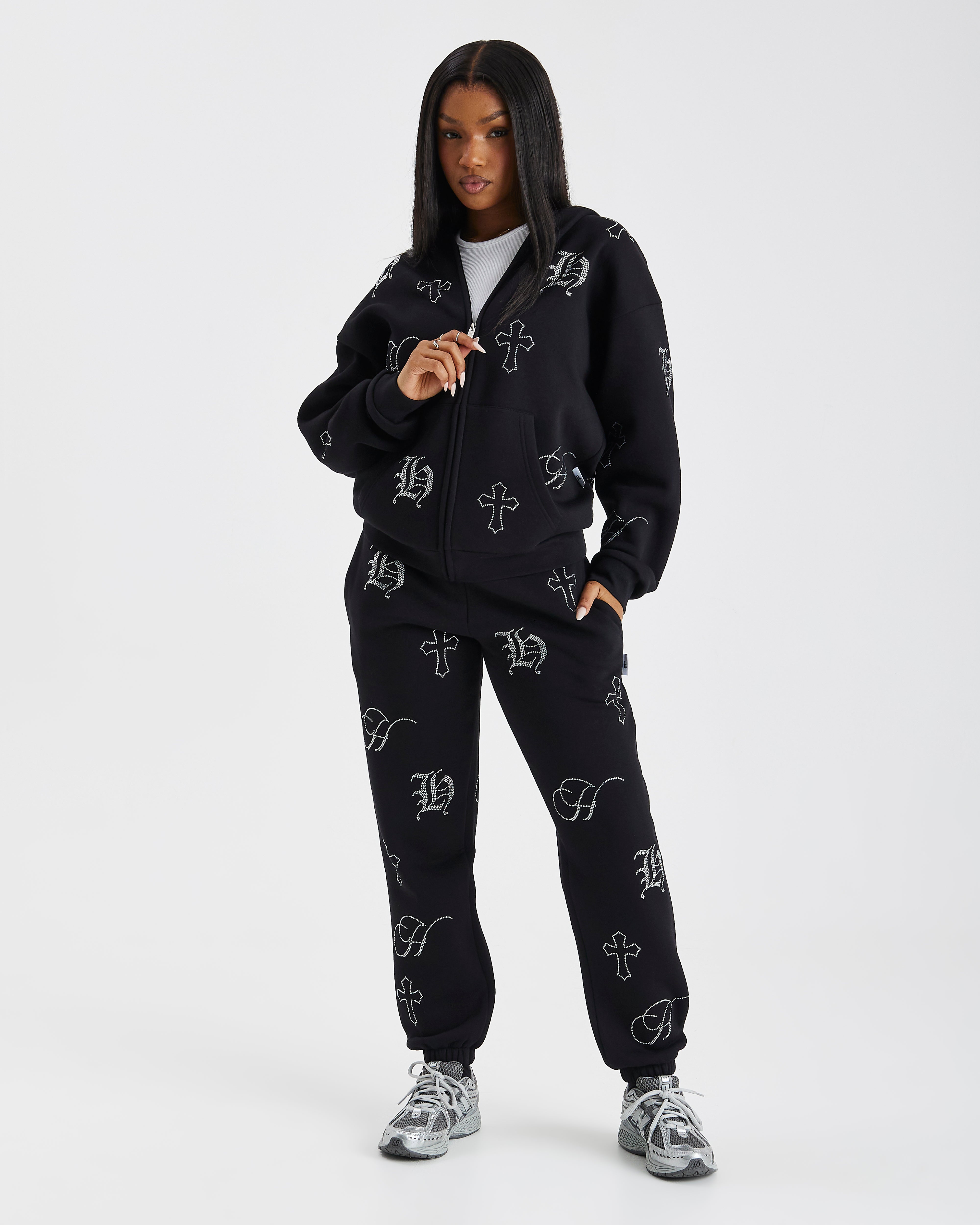 A model wearing a black zip-up hoodie and matching sweatpants set, both featuring a white gothic-style monogram pattern, posed against a light gray studio background.