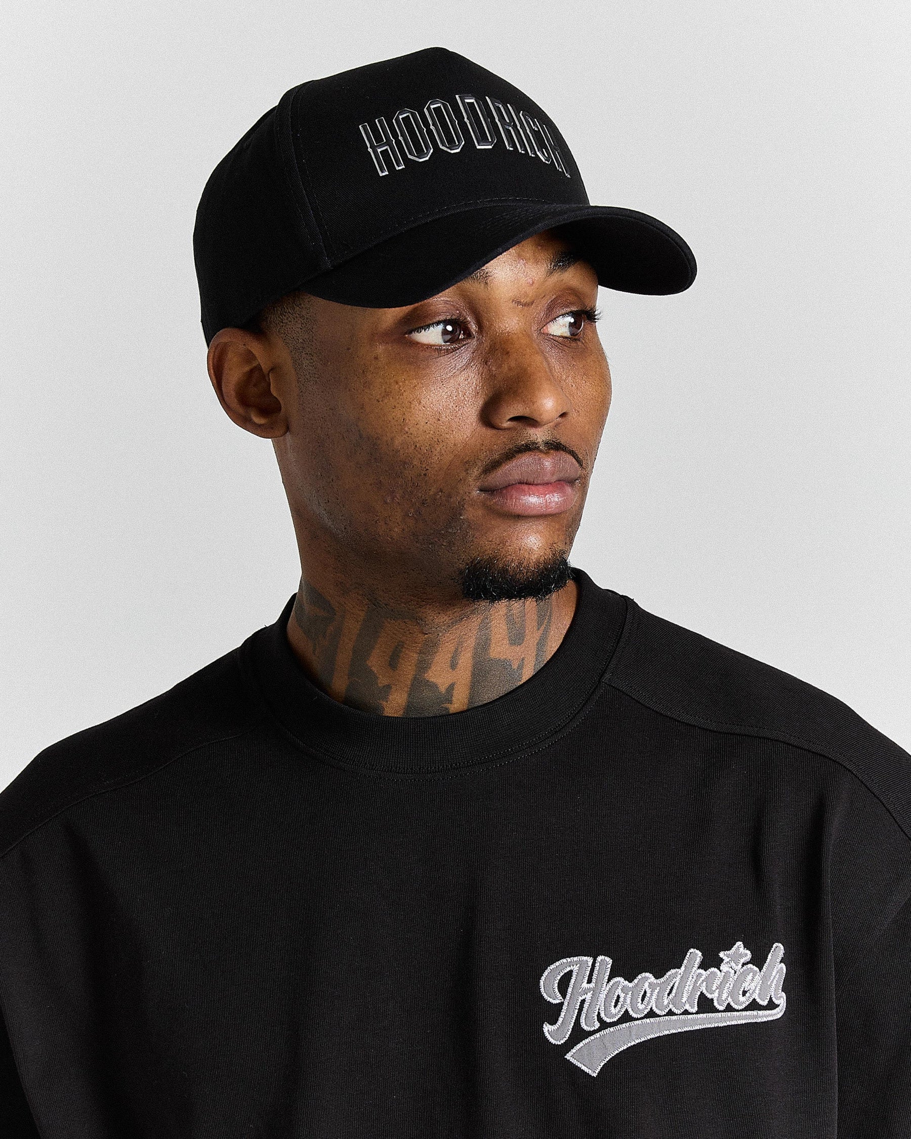 Man wearing a black cap and black sweatshirt with 'Hoodrich' branding on a gray background
