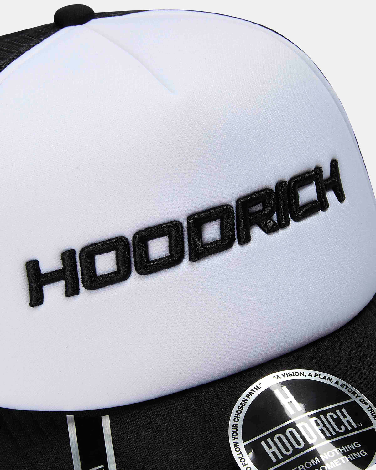 Close-up of a white and black trucker cap featuring black embroidered 'HOODRICH' lettering on the front panel and a black snapback closure.