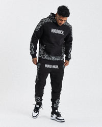 A male model wearing a black joggers with white motif print and text 'HOODRICH' on the hoodie.