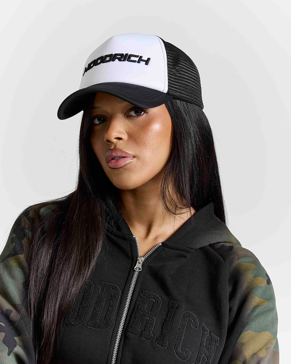 A woman wearing a black and white trucker hat with the brand name 'HOODRICH' on the front, paired with a black zip-up hoodie featuring camouflage sleeves and tonal 'HOODRICH' lettering on the chest.