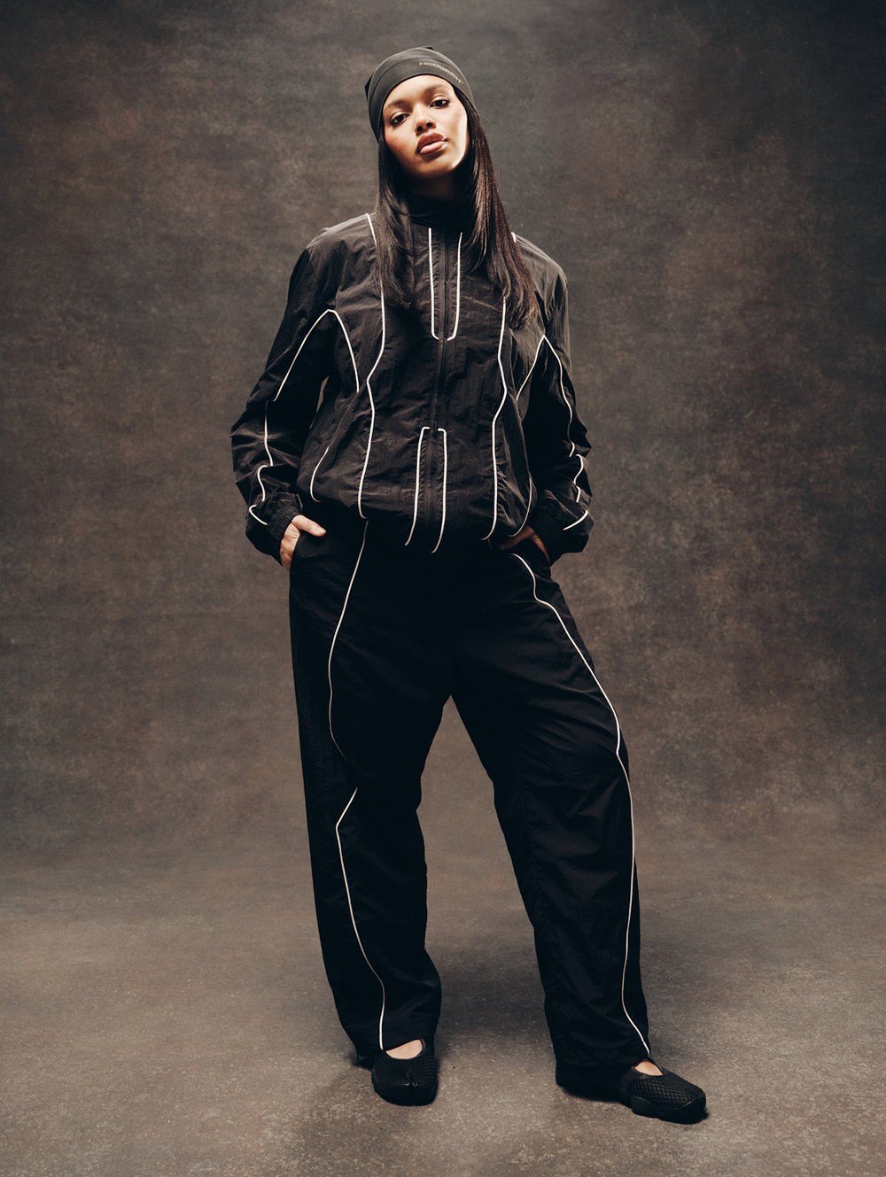 A model wearing a black tracksuit with white contrast stitching, a black beanie, and black flats, standing against a textured dark brown studio backdrop.