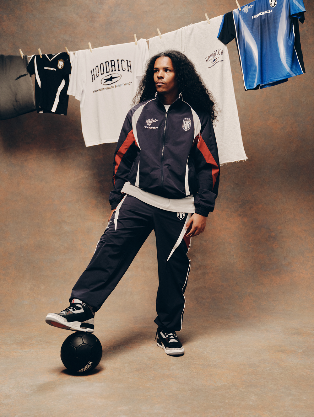 A model wearing a dark navy and white track jacket with red accents and matching track pants stands on a black soccer ball in front of a brown studio backdrop. Sports jerseys are hanging on a clothesline behind them.