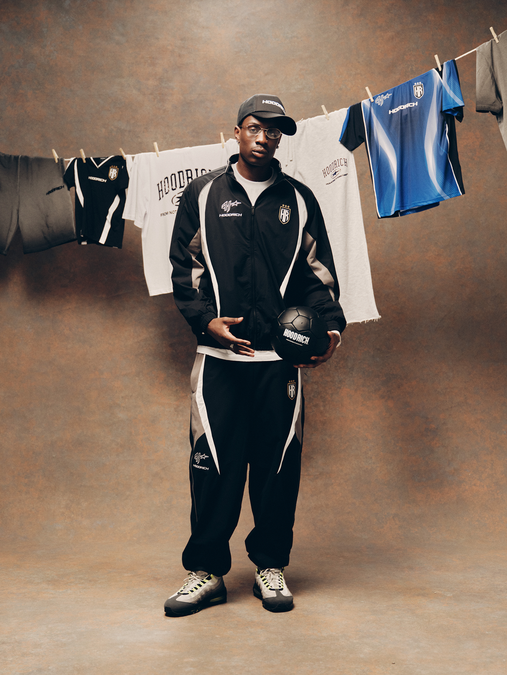 A man stands in a studio wearing a black and white 'HOODRICH' tracksuit with a matching cap, holding a black football. Behind him, a brown textured backdrop features a clothesline with white and blue jerseys hanging on it.
