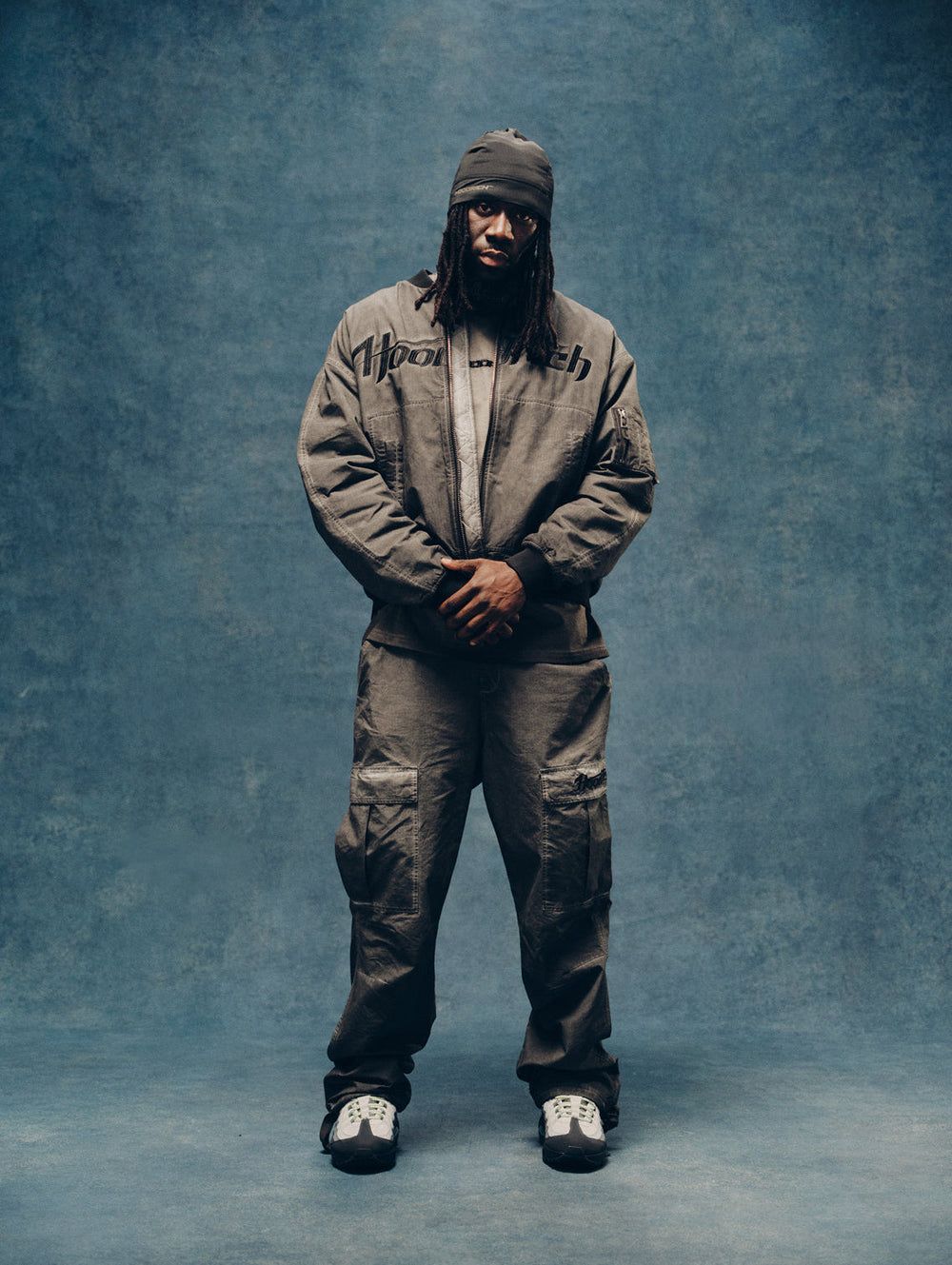 A male model stands against a textured blue studio backdrop wearing an oversized gray zip-front jacket with black 'HOODRICH' script text on the chest, matching gray cargo pants, a dark beanie, and white sneakers.