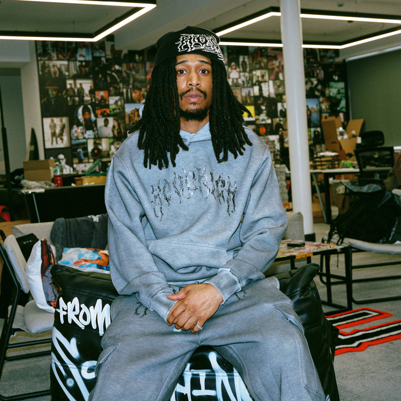 A man with long dreadlocks sits in a black leather chair in a modern office space, wearing a light gray hoodie and sweatpants set with 'hoodrich' printed across the front and matching sweatshirt. He is also wearing a black beanie with a white graphic.