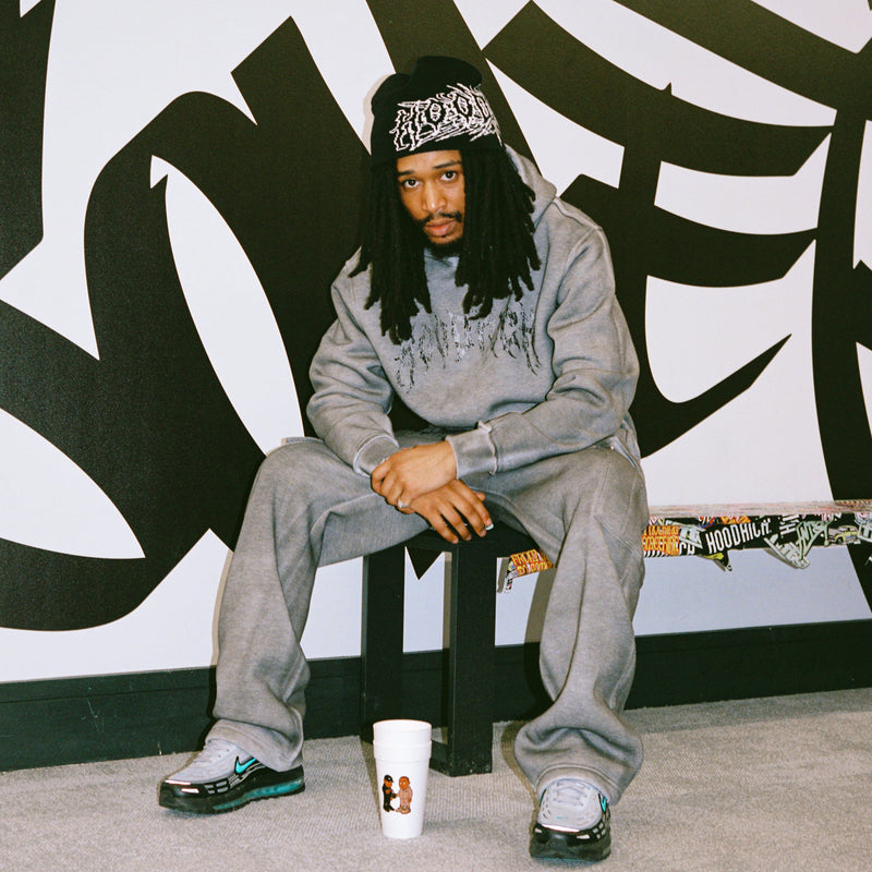 A man with long dreadlocks sits on a black stool in front of a large black graffiti-style mural on a white wall. He is wearing a gray Hoodrich sweatshirt and sweatpants, a black beanie with a white graphic, and sneakers.