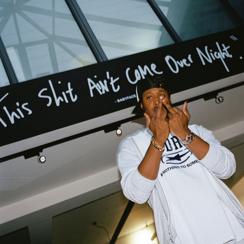 A man wearing a white long-sleeve shirt with a black graphic print and a black cap poses in front of a black banner with white handwritten text.